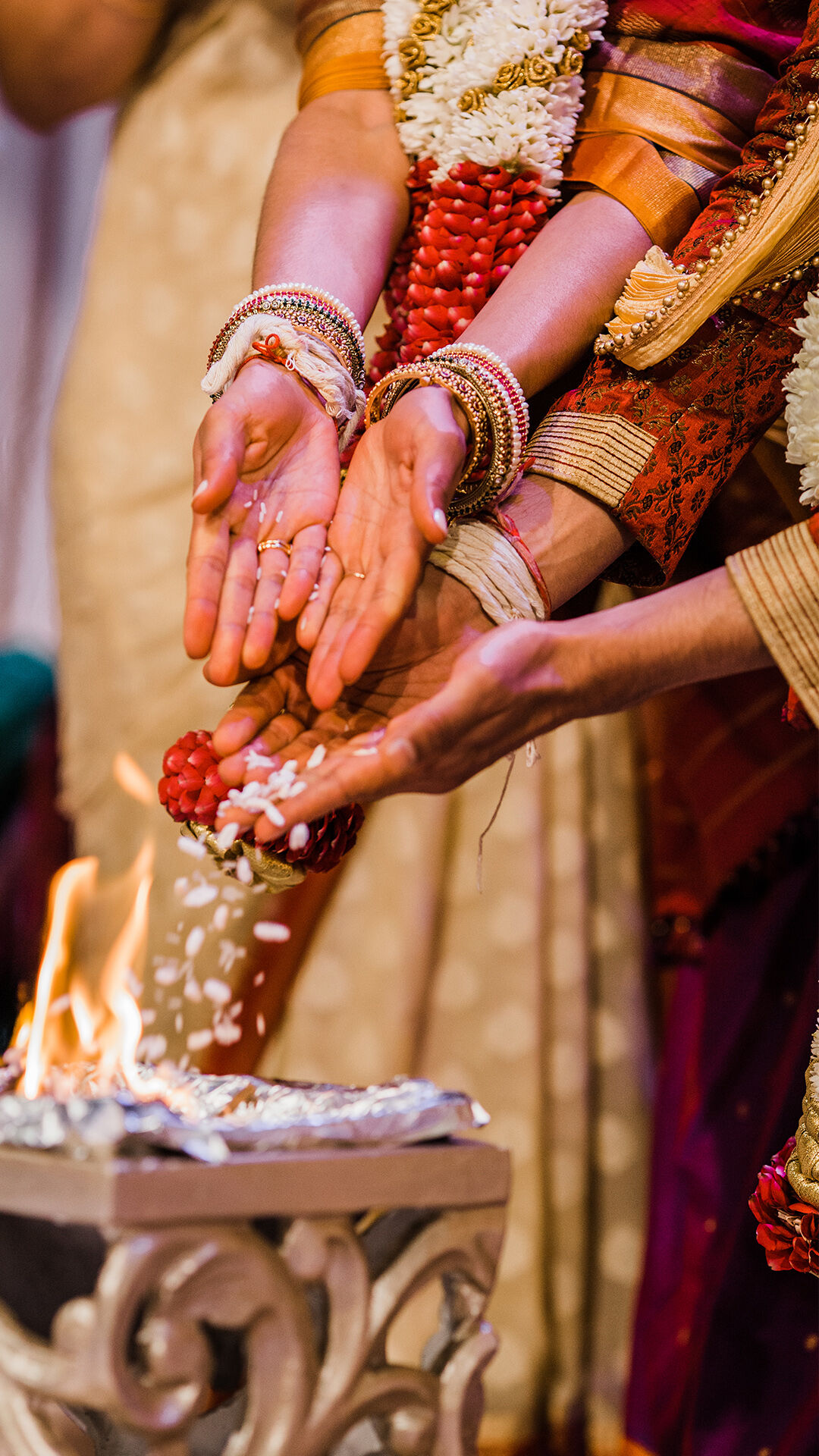 Close-up of a Hindu wedding ceremony where the bride and groom, dressed in traditional attire with garlands, are performing a ritual of offering rice into the sacred fire (Havan).